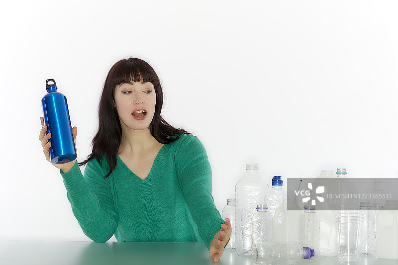 一名年轻女子坐在桌边，手里拿着一个可重复使用的瓶子，推开一堆空塑料瓶图片素材