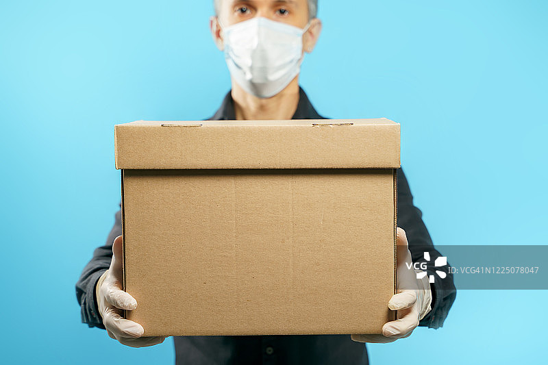 一个纸板箱的特写镜头，在一个年轻男子的手中，在一个医疗防护面具和手套在一个蓝色的背景。安全交付图片素材