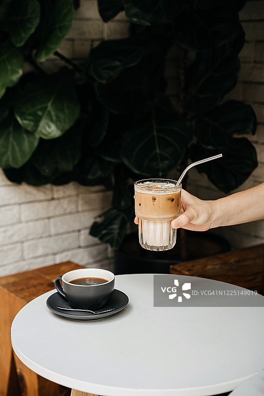 在一家咖啡店的木桌上，一个女人的手拿着一杯冰拿铁和一杯咖啡，靠着白砖墙和盆栽图片素材