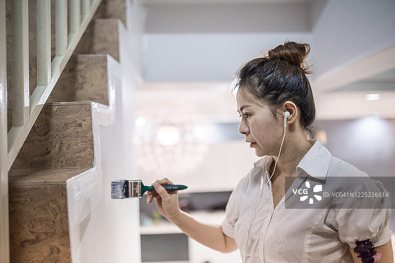 一位亚洲华人妇女在她的新房子里听着音乐，用油漆刷着墙壁。家居改造和装修的概念。图片素材