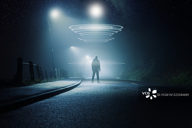 科幻小说中的概念。一个神秘的人站在路中央。在一个有雾的冬天的晚上。明亮的ufo在他头顶发光。图片素材