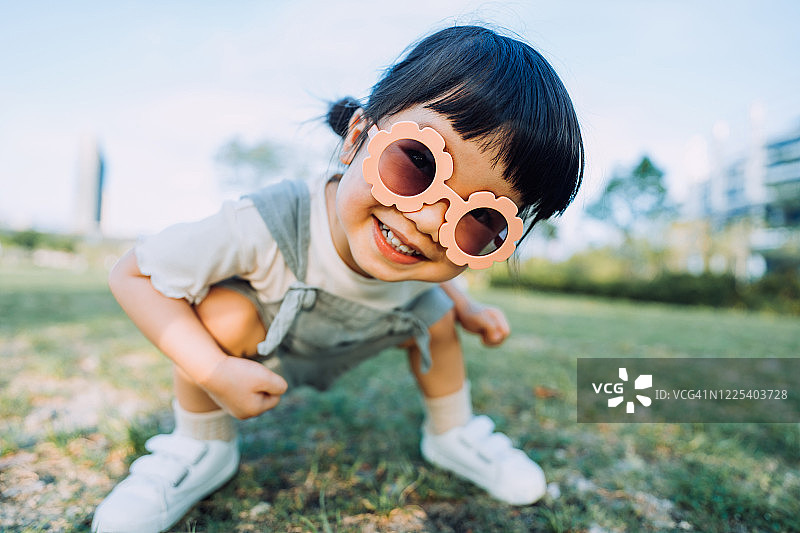快乐的小亚洲学步的女孩与花形太阳镜微笑愉快地在户外享受夏天的日子在公园图片素材