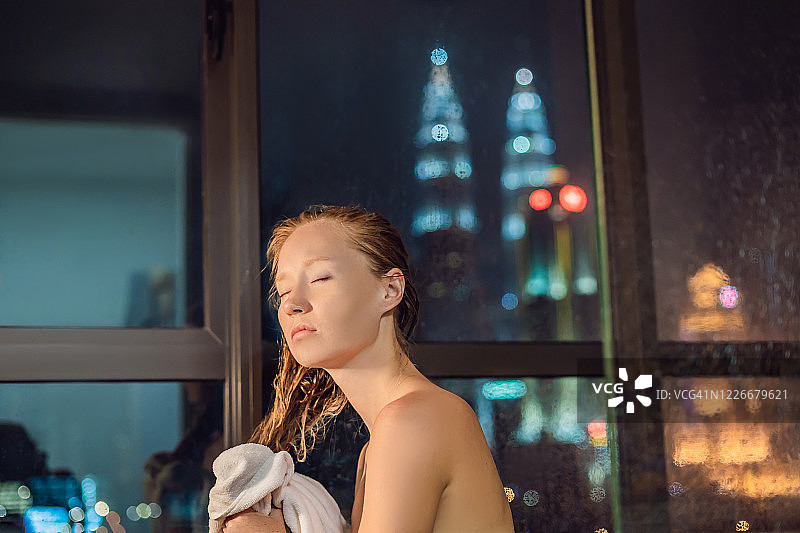 一个美丽的女人在淋浴后擦拭她的头发，在一个窗户的背景，一个夜晚城市的全景图片素材