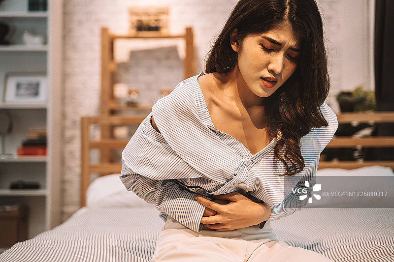 这是一幅20多岁的亚洲年轻女性的肖像，她晚上在卧室里忍受着肚子里的疼痛。腹泻、便秘、肠道、胃炎、月经等症状图片素材