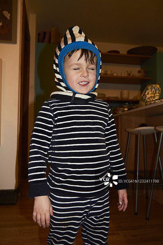 可爱的小男孩穿着蓝白条纹的睡衣和条纹兜帽在他的房子里玩，做鬼脸。他的拖鞋上也有条纹。图片素材