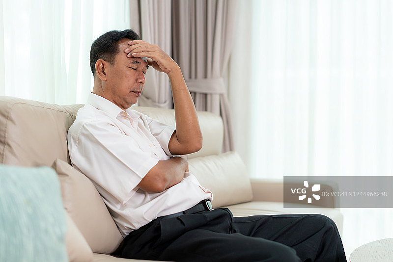 沮丧的亚洲老人感到疲倦和头痛，并覆盖他的头，而坐在客厅的手在家里。图片素材
