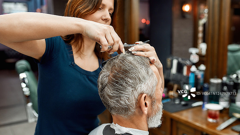 工作过程。一个长着胡子的英俊男子坐在理发店的扶手椅上，年轻的理发师正在为他理发。男子参观美容院图片素材