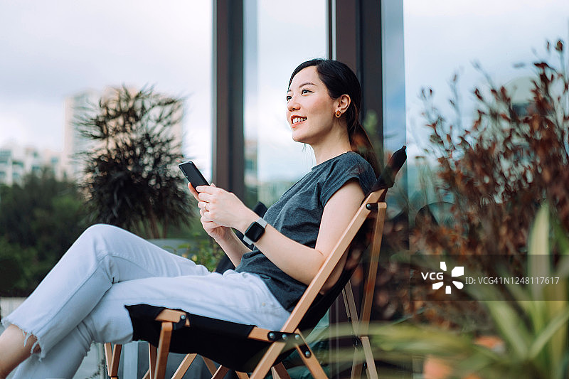 一名欢快的年轻亚洲女子坐在后院的躺椅上，一边用智能手机一边放松，周围是美丽的室内植物图片素材