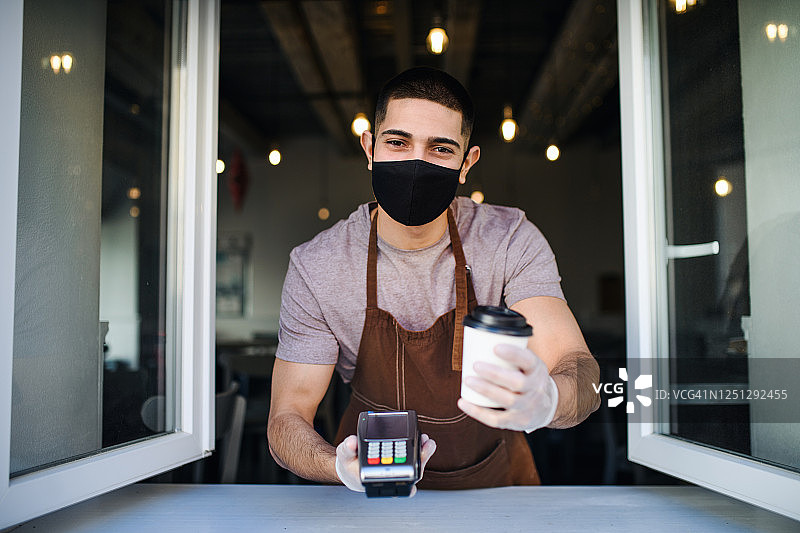 年轻的服务员戴着口罩和手套，通过咖啡店窗口端咖啡，非接触式支付理念。图片素材