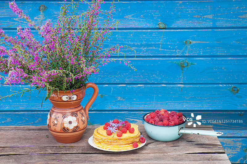 可丽饼配新鲜的覆盆子，紫色的野地花和一碗浆果放在质朴的木桌上图片素材