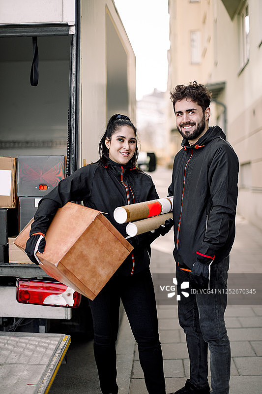 一幅自信的年轻送货男男女女拿着包裹靠着卡车站在城市里的肖像图片素材
