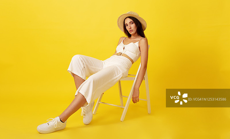 戴草帽的漂亮女人坐在黄色背景的白色椅子上。图片素材