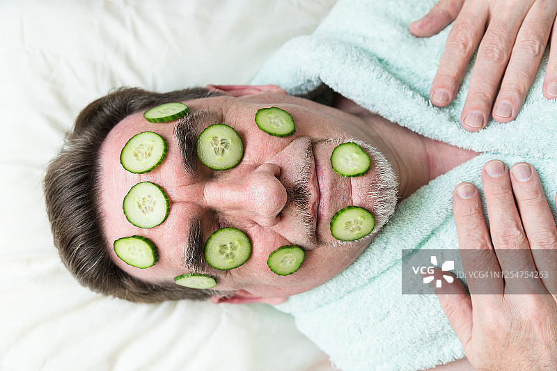 男子的特写照片自制黄瓜面膜在他的脸上。一位老人在隔离期间在家照顾自己的脸。图片素材