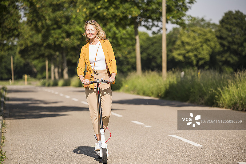 快乐成熟的女人在阳光灿烂的日子里骑电动踏板车在路上图片素材
