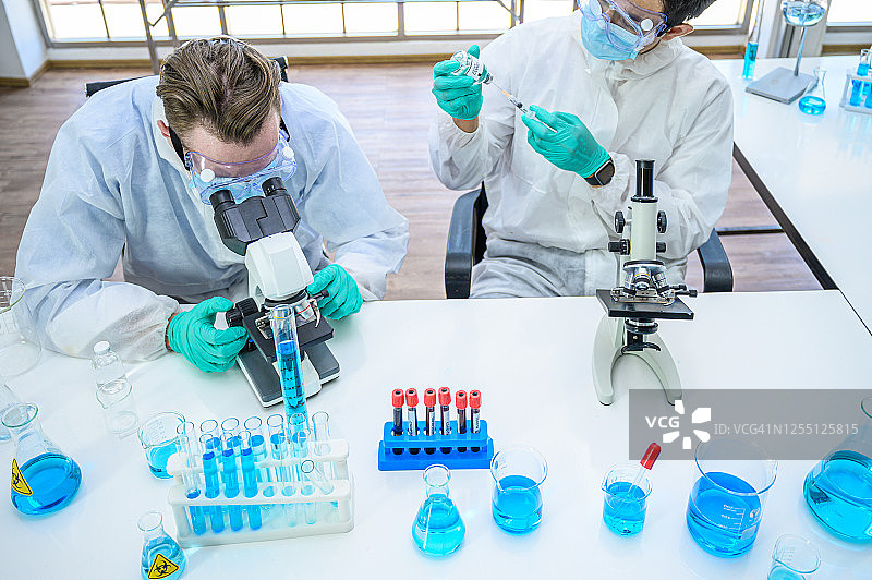 一对男性科学家穿着防护服，在实验室用显微镜和许多实验室设备研究冠状病毒疫苗。图片素材