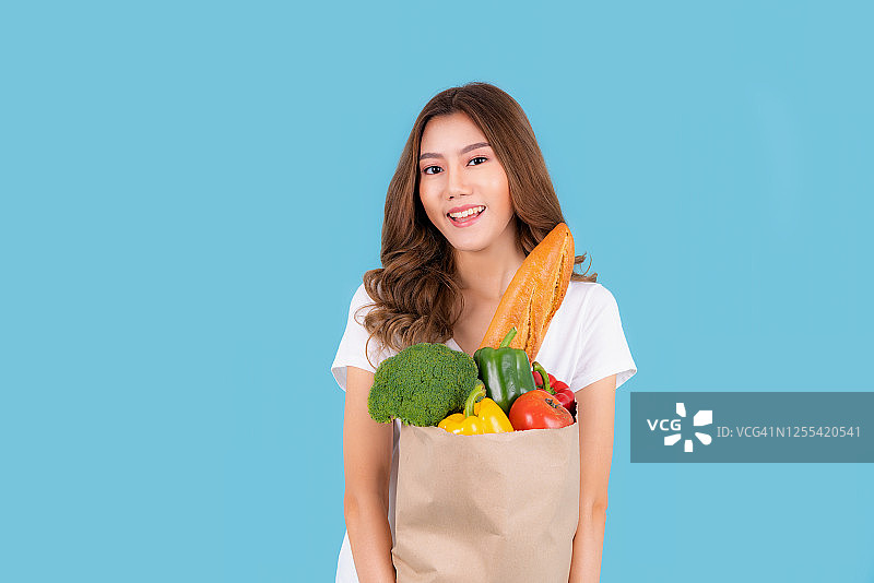 美丽微笑的亚洲女人拿着纸购物袋充满新鲜蔬菜和杂货和微笑，工作室拍摄孤立的青色背景。带一个健康的袋子。图片素材