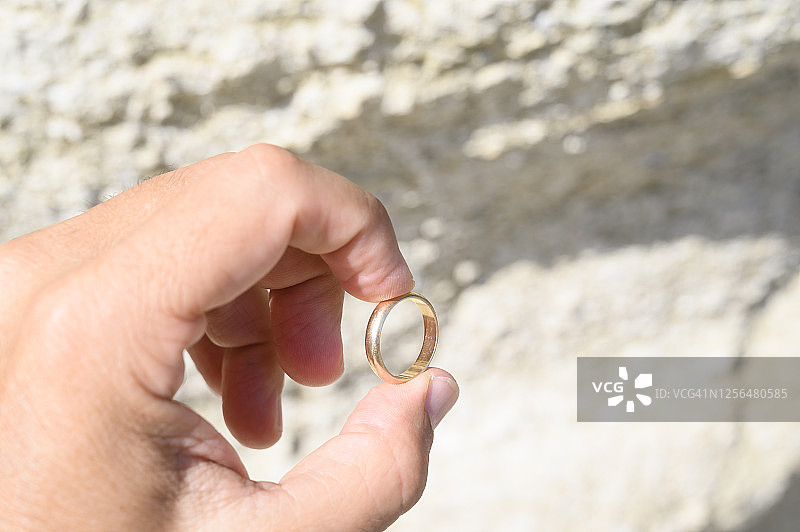 在一个阳光明媚的日子里，一个男人拿着一个岩石背景下的金结婚戒指。图片素材