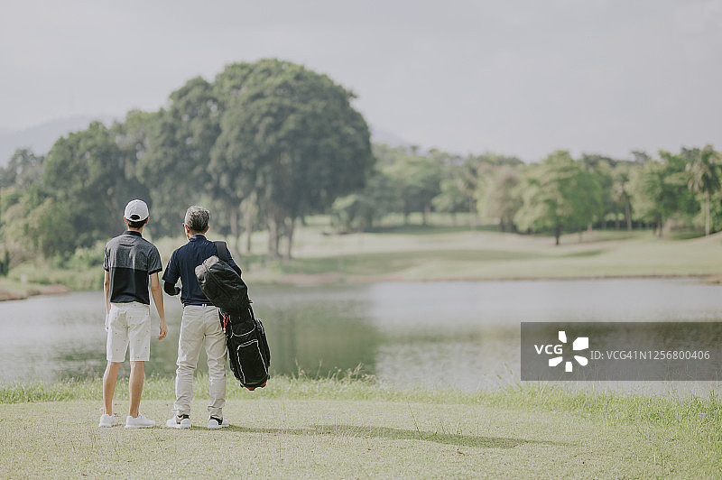 一名亚洲华人男子带着十几岁的儿子往高尔夫球场走去，手里拿着高尔夫球袋看风景图片素材