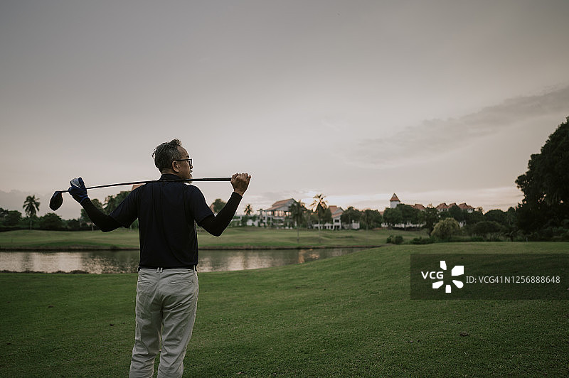 一名亚裔中国资深高尔夫球手肩上扛着高尔夫球杆，看着球场上的景色图片素材
