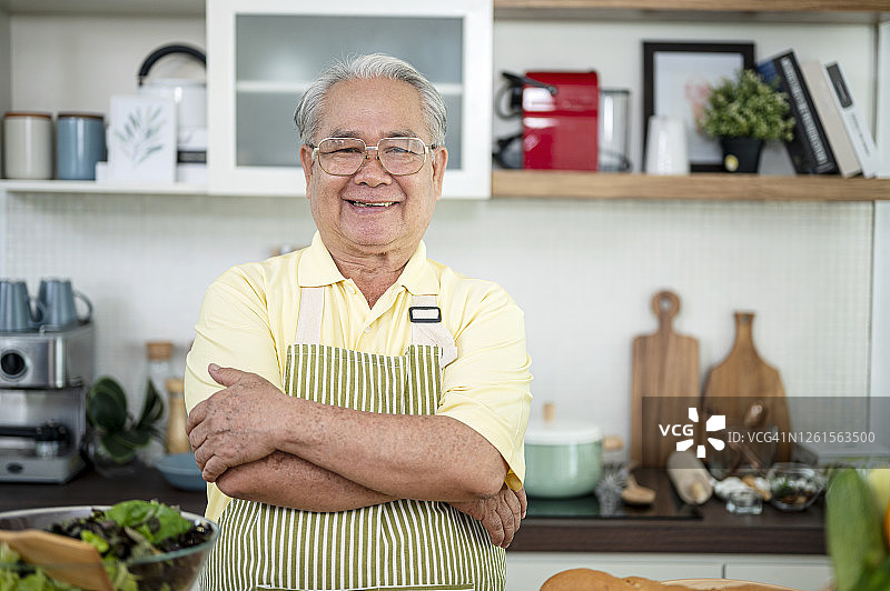 活跃的亚洲资深男子穿着围裙，双臂交叉站在家庭厨房里。准备烹饪，健康的生活方式和烹饪理念。图片素材