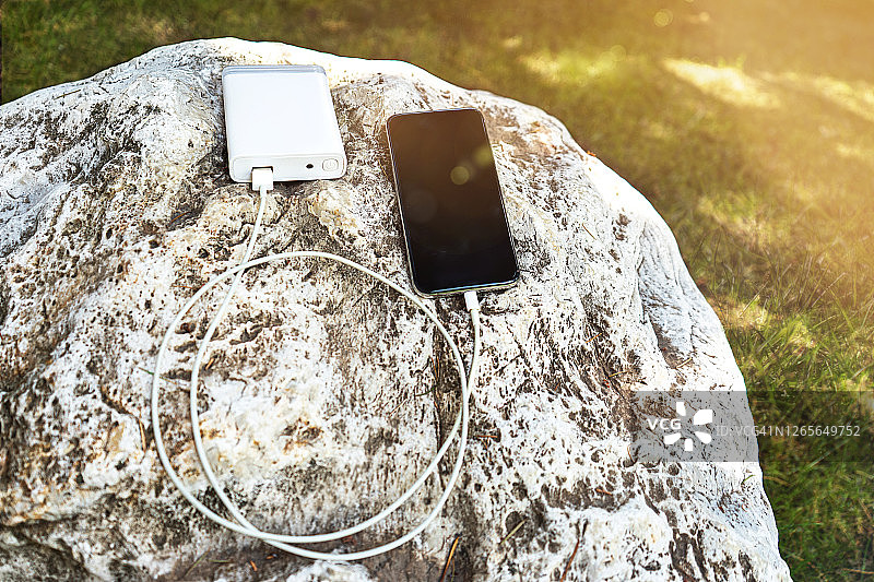 在一个阳光明媚的日子里，在岩石和绿草的背景下，通过设备共享电池无线充电的移动智能手机。图片素材