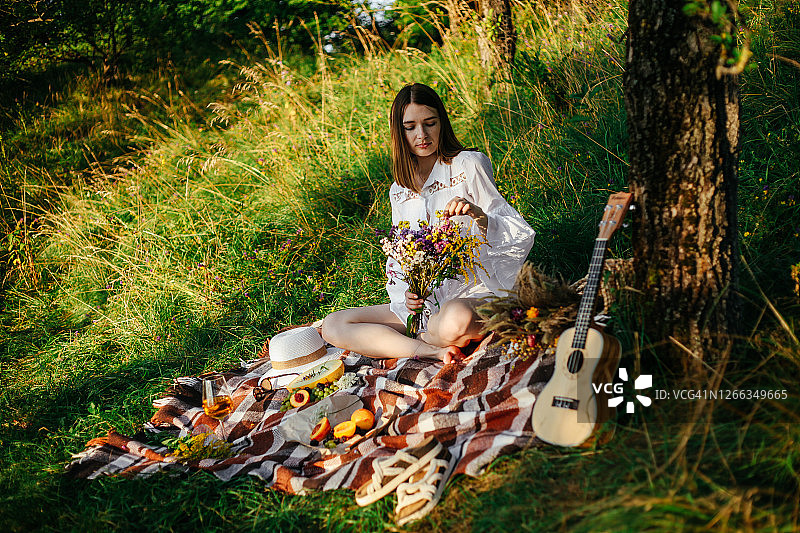 夏季野餐的概念。年轻女子在白色波西米亚风格的连衣裙在夏天的绿色公园有乐趣和放松在阳光灿烂的一天。桌上摆满了水果和浆果，花篮。图片素材
