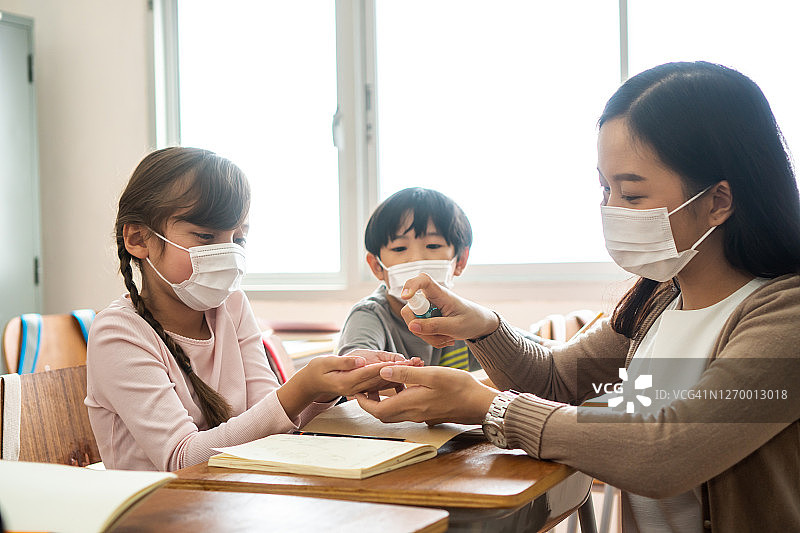 年轻女教师在教室里用酒精喷雾器消毒学生的手。亚洲妇女戴口罩，用洗手液清洁学生的手。学校在隔离和封锁后重新开学图片素材