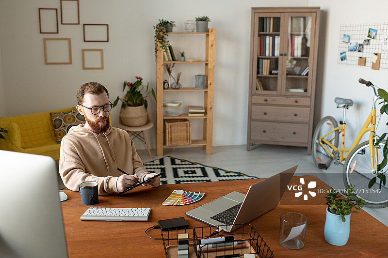 年轻的留着胡子的自由设计师使用平板电脑，触控笔，笔记本电脑和调色板图片素材