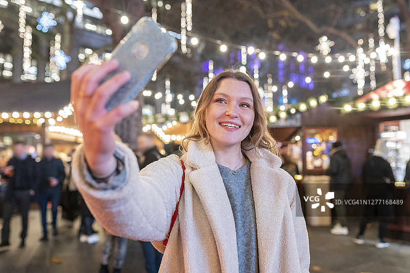 一名年轻女子站在灯火辉煌的圣诞市场夜市上自拍图片素材