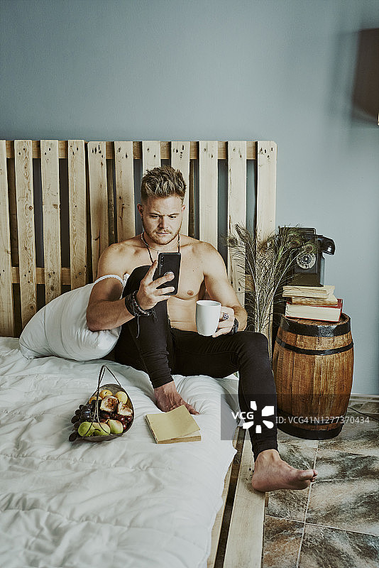 一个年轻人坐在床上，边喝咖啡边玩手机图片素材