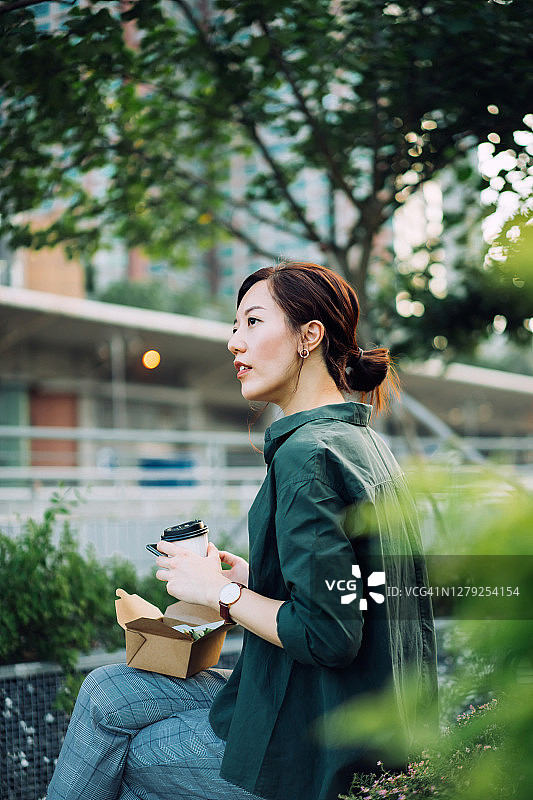 年轻的亚洲女商人在城市公园里的午餐休息时间一边用智能手机一边吃着健康的沙拉午餐盒和一杯咖啡图片素材