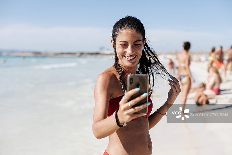 微笑的女人穿着比基尼自拍与手机在海滩对着天空图片素材
