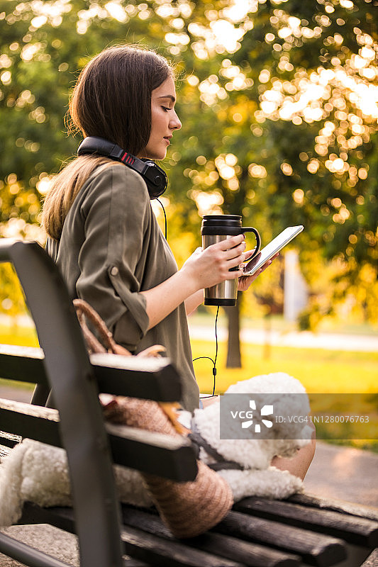 一个年轻女子在公园的长椅上一边玩手机一边喝咖啡图片素材