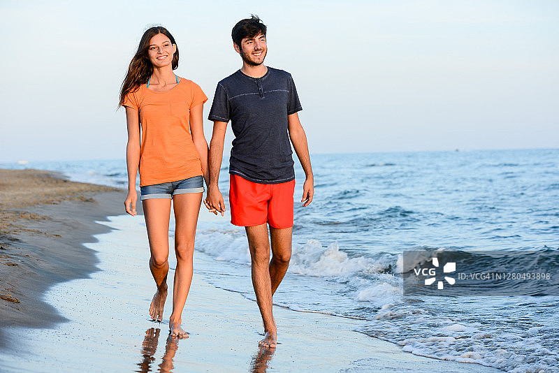 一对浪漫的年轻情侣在日落时分一起在地中海海滩的沙滩上赤脚散步图片素材