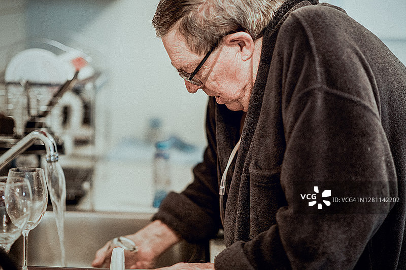 一位老人站在他的厨房里，正在为他每日的支气管扩张治疗准备他的吸入器。图片素材