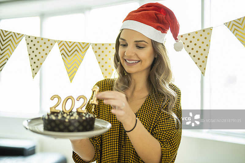 一名年轻女子在家庆祝圣诞节，手里拿着巧克力蛋糕，蛋糕上点燃了数字2020形状的蜡烛图片素材