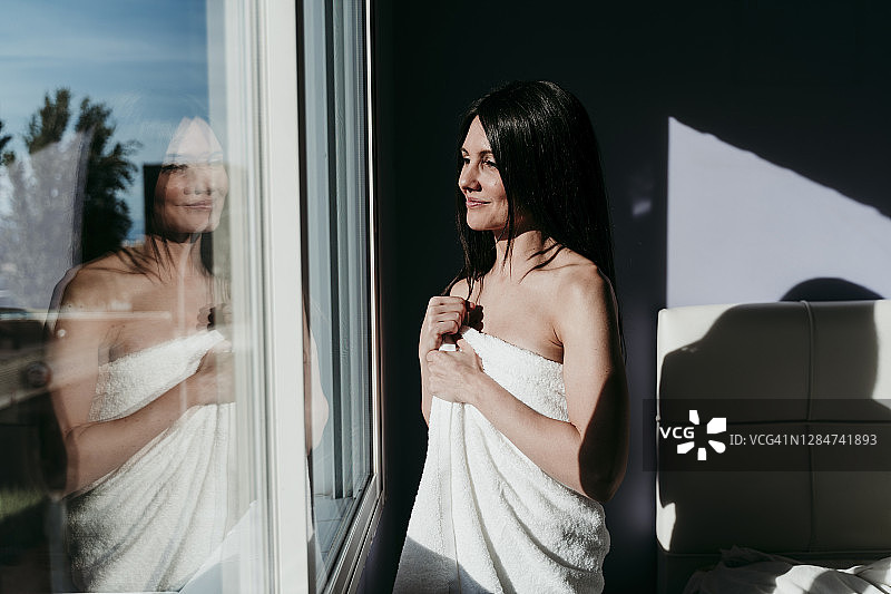 微笑体贴的女人裹着毛巾站在家里的窗口图片素材