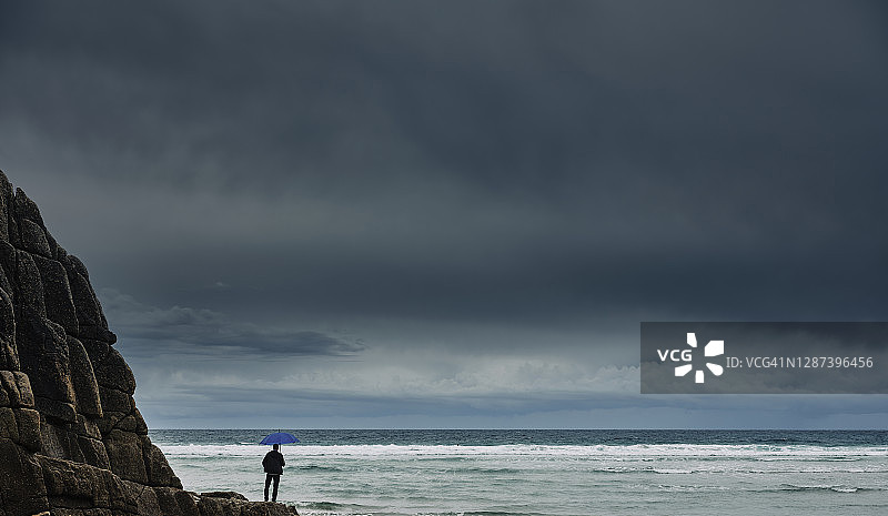 人们撑着伞站在海边的岩石上，乌云密布，远处天空更清澈。图片素材