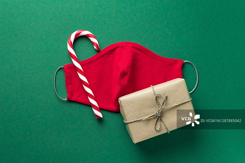红色医用防护口罩，拐杖状条纹糖果，绿色圣诞背景的礼物。预防和预防COVID - 19大流行的传播。一个快乐健康的圣诞节的概念。图片素材
