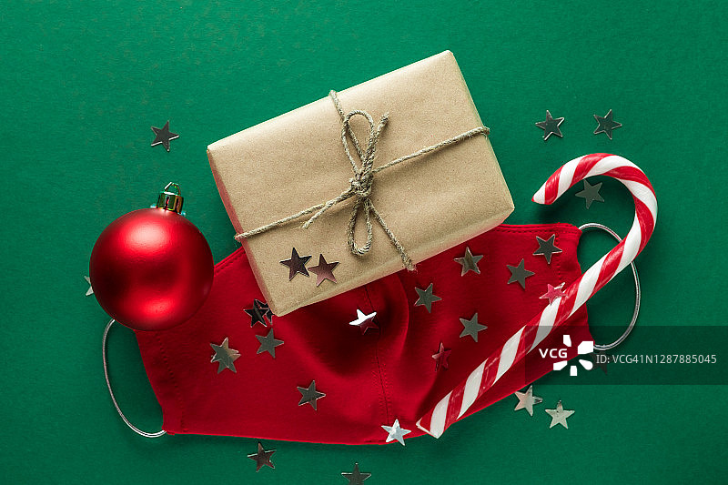 红色医用防护口罩，条纹拐杖糖，圣诞树的礼物和玩具，绿色背景上星星形状的五彩纸屑。预防和预防COVID - 19大流行的传播。快乐健康圣诞的理念。图片素材