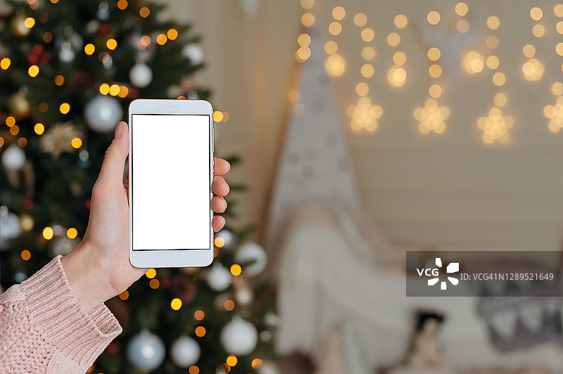 在圣诞装饰圣诞树的背景上，女孩手中的智能手机的模型。图片素材