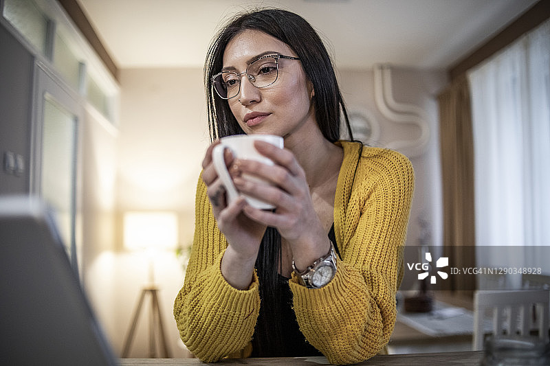 一位年轻女子在家里用一台电脑和一部智能手机工作。一位穿着漂亮黄色毛衣的黑发美女坐在家里，用智能设备在网上工作图片素材