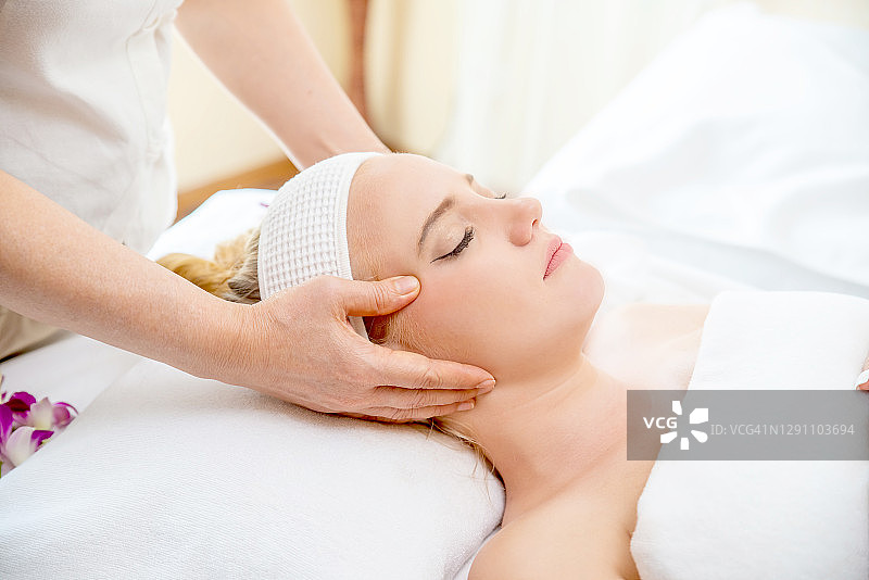 年轻美丽的白种女子躺在spa床上接受美容院按摩师的面部按摩和精油护肤。健康身体按摩和脸部水疗概念图片素材