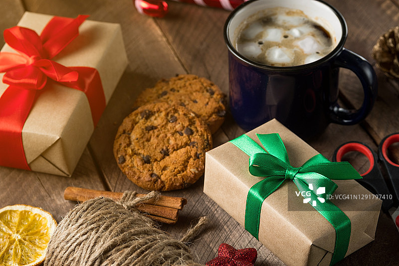 准备一个家庭假日的概念，快乐的平安夜。用亮绿色的丝带把圣诞礼物和带蝴蝶结的盒子捆好，放在木桌的背景上。一杯热咖啡，棉花糖，饼干和巧克力。图片素材