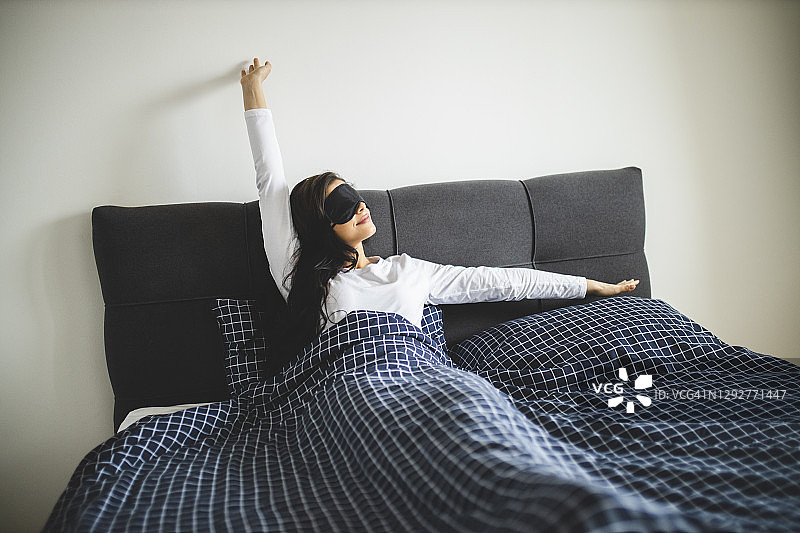 年轻女子戴着睡眠面罩，坐在床上伸展身体图片素材