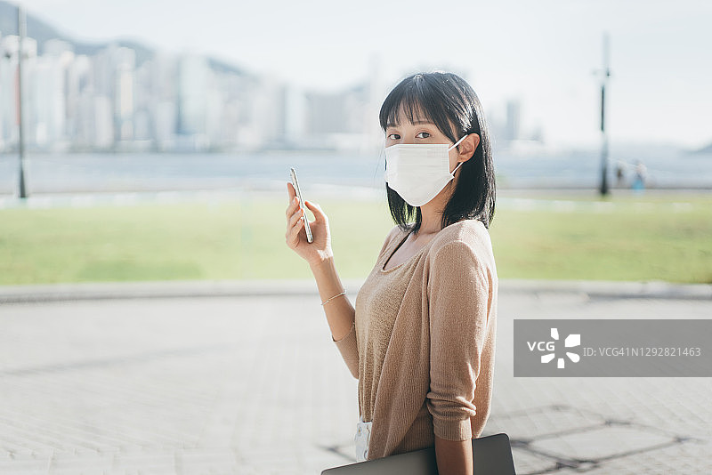一名年轻的亚洲女性戴着防护口罩，以保护和防止病毒在城市里传播，并一边盯着手机和笔记本电脑，一边看摄像头。图片素材