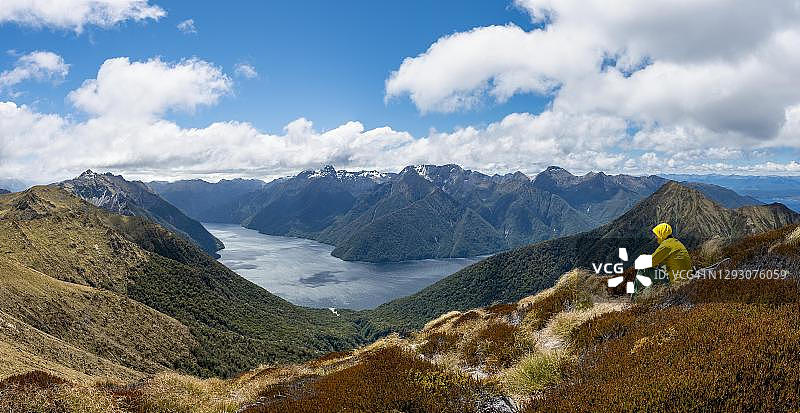 在新西兰南岸峡湾国家公园的开普勒步道上，一名登山者，徒步旅行者坐在草地上，背景是特阿瑙湖的南峡湾，默奇森山脉和南阿尔卑斯山图片素材