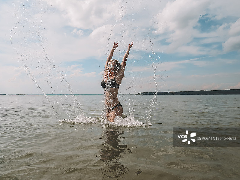 美丽的年轻女子在一个炎热的夏天，穿着黑色比基尼，水溅在海滩浅水的背景图片素材