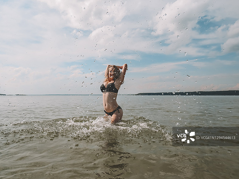 美丽的年轻女子在一个炎热的夏天，穿着黑色比基尼，水溅在海滩浅水的背景图片素材
