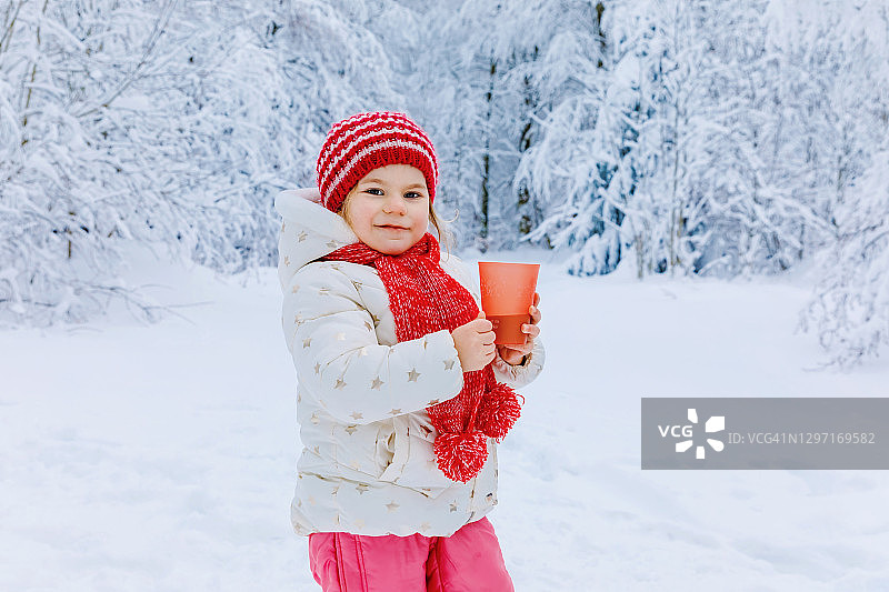 可爱的蹒跚学步的女孩喝热巧克力在冬天的森林。快乐健康的孩子与一杯热气腾腾的可可或茶，户外。33、活跃家庭休闲，漫步雪景。图片素材